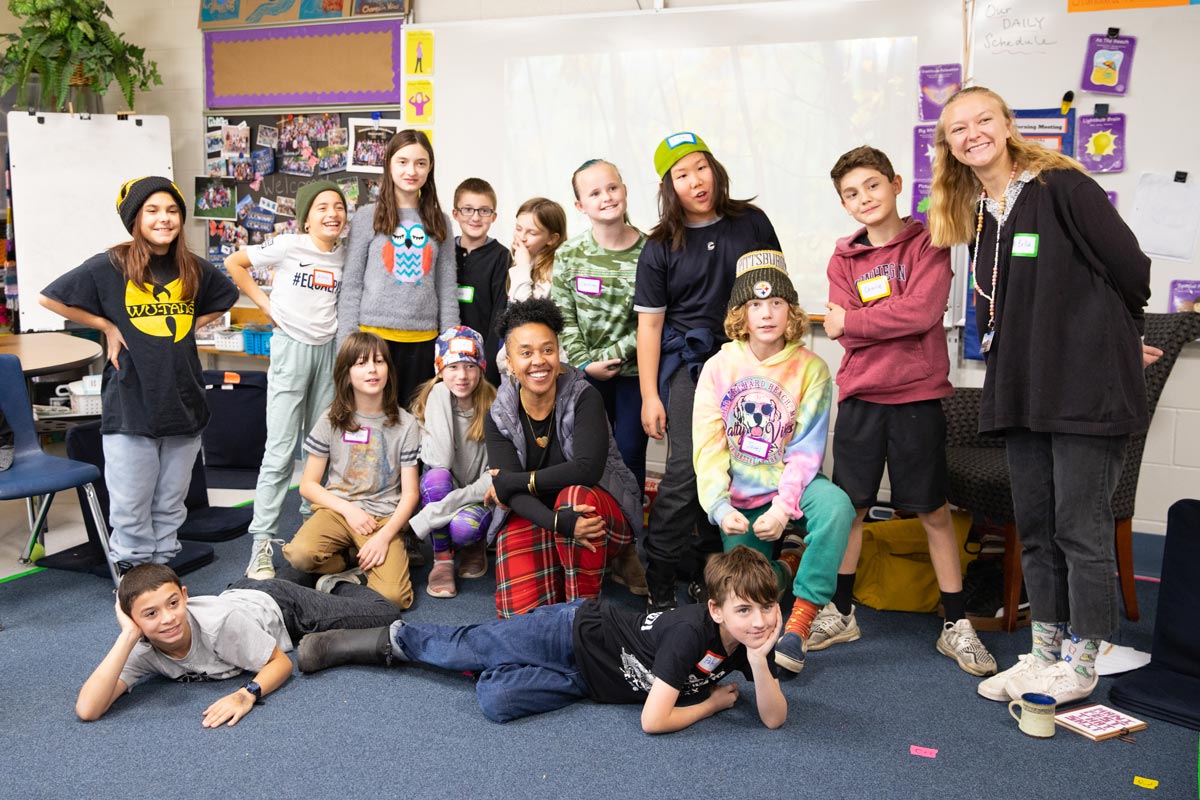 A group of students and two adults gather in a colorful classroom, some standing and others sitting or reclining on the floor smiling for the photo.