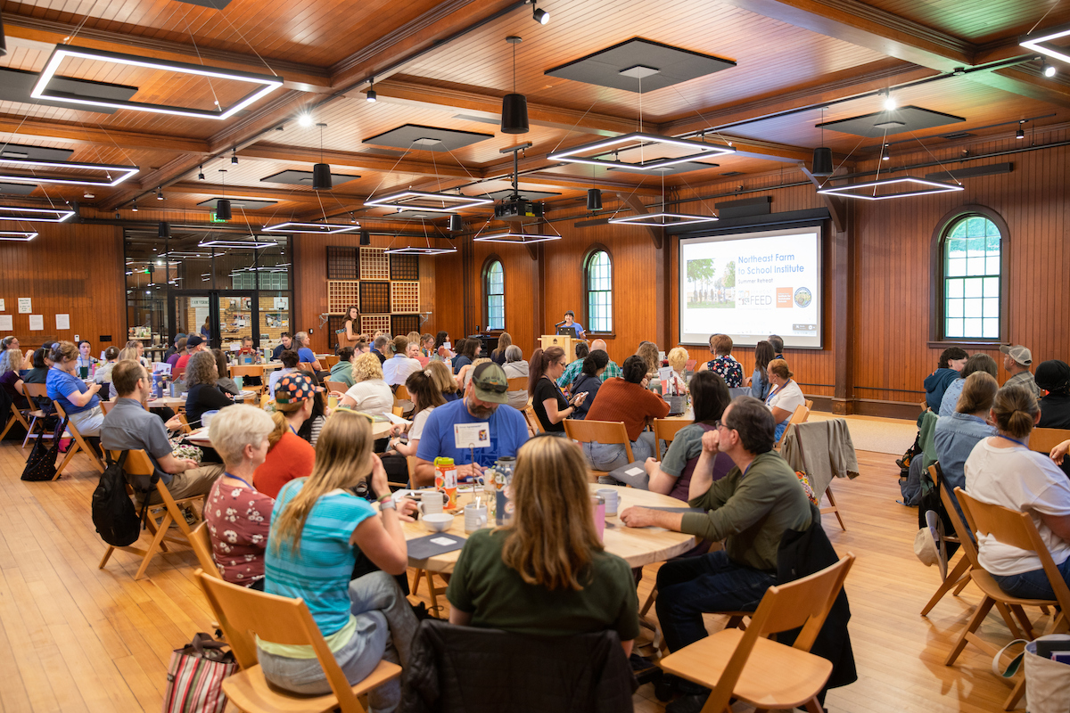 A large group of people sits at round tables in a spacious room in the Coach Barn. They face a presentation screen displaying "Northeast Farm to School Institute."