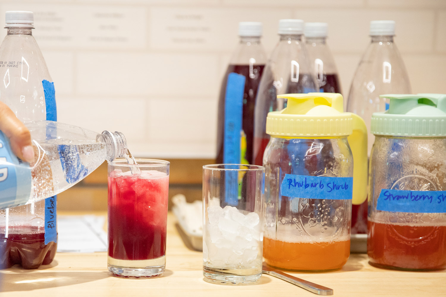 A variety of colorful drinks are displayed on a countertop. Bottles labeled "Rhubarb Shrub" and "Strawberry Shrub" are visible, along with jars of other mixtures.