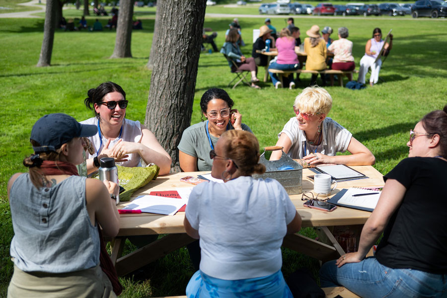 A group of people sit around a picnic table engaging in conversation. Other groups are visible in the background.