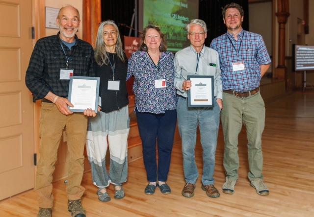 a group of 5 people (2 women; three men) smile at camera. Two of the men are holding award plaques.