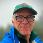 Man in black glasses and a green hat smiles in front of a blurred green background.