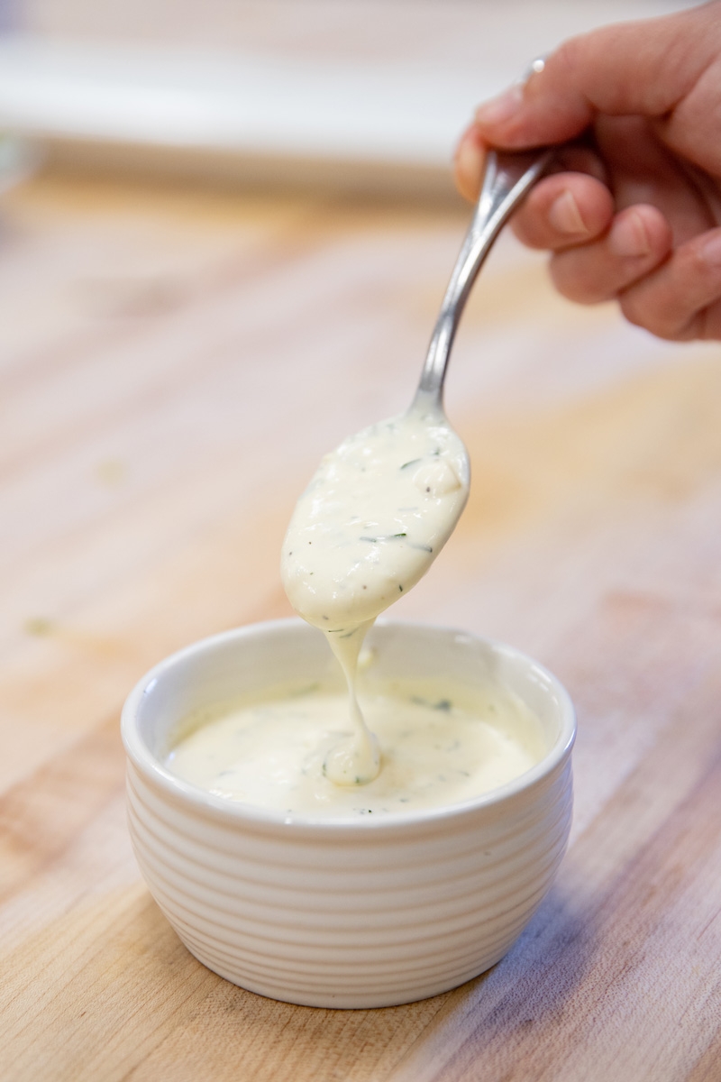 A hand holding a spoon with a portion of creamy, herb-infused sauce above a small, white ramekin filled with the same sauce. 