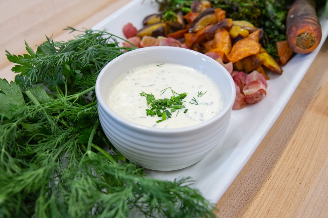 A white bowl of creamy dressing garnished with fresh herbs, placed next to a colorful assortment of roasted vegetables, and fresh dill on a platter.